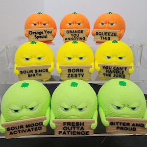 Pode incluir: Uma coleção de nove figuras de frutas antropomórficas. As figuras laranja, amarelas e verdes seguram cada uma uma placa com uma frase engraçada. As figuras laranja têm as frases "Orange You Special", "Orange You Annoying" e "Squeeze This".