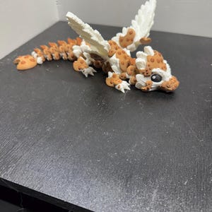 May include: A 3D-printed articulated dragon toy. The dragon features a white body with brown spots, orange accents, and white wings. The dragon is lying on a dark surface.