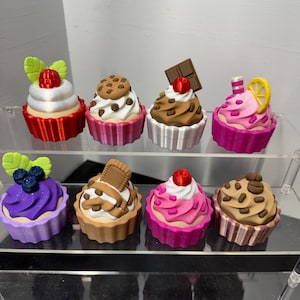 May include: A collection of eight colorful cupcake figurines with various toppings, including berries, cookies, and chocolate. Each cupcake has a unique design and sits in a decorative, fluted cup. The cupcakes are arranged on a clear display shelf.