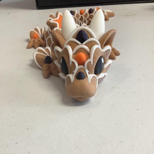 Puede incluir: Una figura de dragón marrón y blanco con detalles en naranja y negro. El dragón tiene grandes cuernos blancos, ojos negros y un patrón de escamas detallado. La figura está hecha de un material que se asemeja al pan de jengibre.