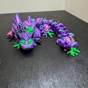 May include: A purple articulated dragon figurine with green, pink, and black accents. The dragon has a detailed head and body, with a segmented design allowing for flexible posing. The figurine is made of plastic and sits on a dark surface.