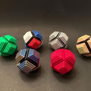 Puede incluir: Siete bolas de rompecabezas geométricas multicolores están dispuestas sobre una superficie oscura. Las bolas son de varios colores, incluyendo verde, rojo, azul, plateado y beige. Cada bola tiene una superficie texturizada y tachonada y un diseño segmentado único.