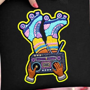 Puede incluir: Bolso tote negro con un diseño bordado colorido. El diseño incluye patines, un radiocasete y manos. Los patines son azules, rosas y amarillos. El radiocasete es negro y morado. Las manos son marrones.