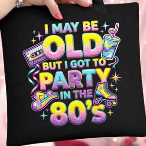 Puede incluir: Bolso tote negro con un diseño colorido de los años 80. El texto dice "I MAY BE OLD BUT I GOT TO PARTY IN THE 80'S" con imágenes de un casete, un patín y un vaso.