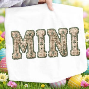 May include: A white tote bag with the word "MINI" embroidered in a woven, textured style. The letters are beige with a green border and small white bead accents. The background features grass, flowers, and Easter eggs.