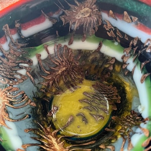 May include: A decorative glass bowl with a vibrant, layered design. The rim features bands of red, black, white, and green. The interior has a yellow center and is adorned with intricate, brown, spiky, organic shapes, creating a unique artistic piece.