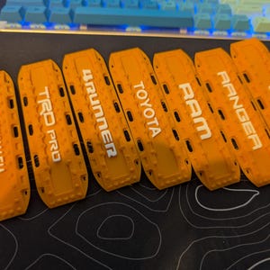 May include: Seven orange recovery boards with white lettering. The boards display the following text: TOYOMAFIA, TRD PRO, 4RUNNER, TOYOTA, RAM, RANGER, and Jeep. The boards are arranged on a black surface.
