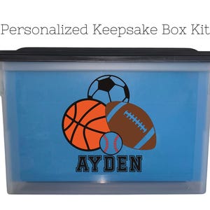 May include: A clear plastic storage box with a black lid. The box has a blue background with a design of a basketball, football, soccer ball, and baseball. The name "AYDEN" is printed in black letters below the balls.