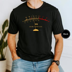 May include: Black t-shirt featuring a VU meter graphic with the text "DECIBEL POWER LEVEL". The meter has yellow and red accents, with numbers indicating decibel levels. The shirt is a classic crew neck.