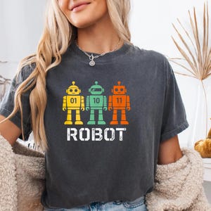 Peut inclure: T-shirt gris foncé avec trois robots colorés. Chaque robot est jaune, vert ou orange et porte un numéro : 01, 10 et 11. Le mot "ROBOT" est imprimé en blanc en dessous. Le t-shirt est de style décontracté.