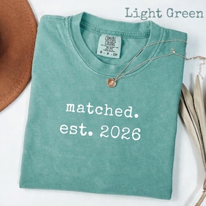 May include: Light green Comfort Colors t-shirt with the text "matched. est. 2026" in white. A silver necklace with a small pendant rests on the shirt. The shirt is folded, and a brown hat is visible in the background.