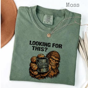 Könnte beinhalten: Moosgrünes T-Shirt mit dem Text "LOOKING FOR THIS?" und einer Grafik eines lächelnden Bigfoot, der eine 10-mm-Stecknuss hält. Eine silberne Halskette mit Anhänger befindet sich auf dem Shirt.
