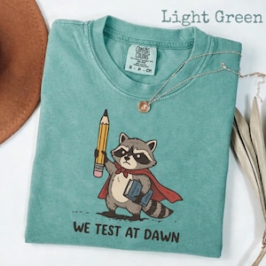 May include: Light green t-shirt with a graphic of a raccoon wearing a cape and holding a pencil and books. The text "WE TEST AT DAWN" is printed below the raccoon. A silver necklace with a pendant is draped on the shirt.