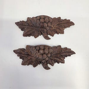 May include: Two brown, carved wooden grape and leaf decorations. Each piece features a cluster of grapes and detailed leaves, with a rustic, aged appearance. The decorations are likely for home decor or craft projects.