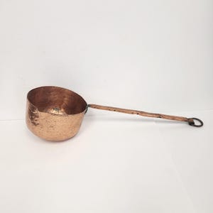 May include: A hammered copper ladle with a long handle and a ring on the end. The ladle is a traditional kitchen tool used for scooping liquids.