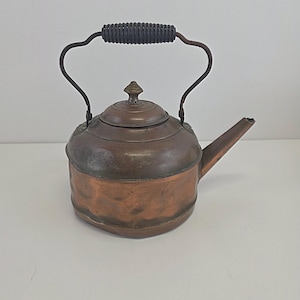May include: A copper kettle with a dark handle and spout. The kettle has a round body with a lid and a decorative finial. The handle is made of a dark, coiled material. The kettle is likely used for boiling water.