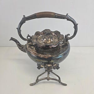 May include: Antique silver-plated teapot with a decorative handle and spout. The teapot has an ornate design on the lid and sits on a three-legged stand. The handle features a brown wooden grip.