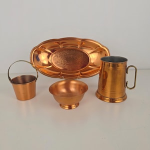 May include: A set of copper kitchen items: a small bucket with a handle, an oval tray, a small bowl, and a large mug with a handle. All items are made of shiny copper, reflecting the light.