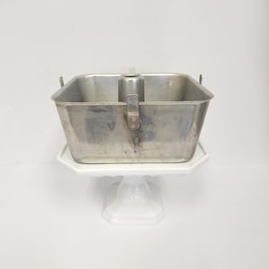 Może przedstawiać: Srebrna metalowa forma do ciasta bundt z wyjmowanym środkowym rurem. Forma stoi na białym stojaku na ciasto.