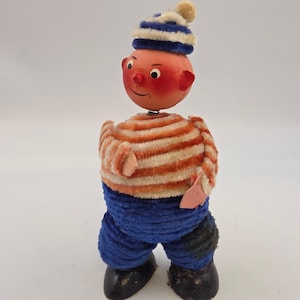Puede incluir: Una figura de juguete vintage con una cara pintada, mejillas rojas y una camisa a rayas naranja y blanca. La figura lleva un gorro a rayas azul y blanco y pantalones azules. El juguete tiene zapatos negros y un resorte que conecta la cabeza y el torso.