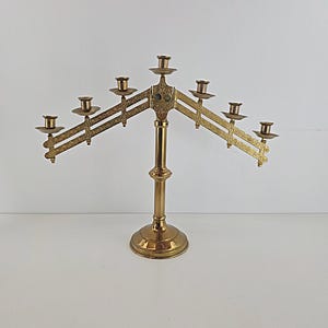 May include: A polished brass candelabra featuring a central vertical stand and two adjustable, hinged arms that extend outward. The arms hold a total of seven candle cups, each with a small circular drip plate, and feature an intricate, embossed pattern.