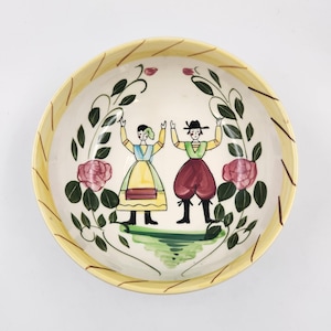 Puede incluir: Un tazón de cerámica blanca con un borde amarillo y un diseño pintado a mano de dos figuras con ropa tradicional bailando en un campo verde con rosas rosas y hojas verdes.