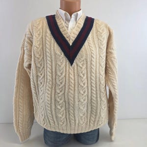 May include: A cream-colored cable knit sweater featuring a V-neckline with dark navy blue and maroon striped trim. The long-sleeved pullover is worn over a white collared shirt and is displayed on a mannequin torso with blue denim jeans.