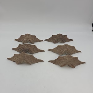 May include: Six brown, leaf-shaped decorative accents are arranged on a white surface. Each accent has a detailed, textured surface with visible veins, and a slightly curved shape. The leaves are evenly spaced.
