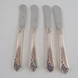May include: A set of four silver-toned butter knives arranged vertically. Each knife features a smooth, flat handle with intricate embossed floral and leaf detailing at the base and a curved, polished metal blade for spreading butter or soft foods.