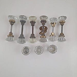 May include: A collection of six glass door knobs with fluted designs and metallic bases, including five complete sets with spindles and three loose knobs. The hardware features clear glass, brass, and dark metal finishes in various states of wear.