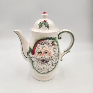 May include: White ceramic teapot with a green handle and a Santa Claus face on the front. The lid has a green holly design.