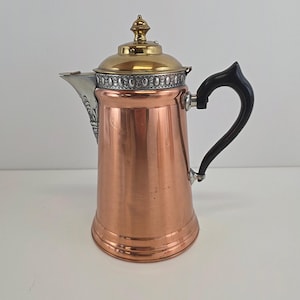 May include: A copper coffee pot with a brass lid and a black handle. The pot has a silver spout and decorative trim around the lid. The pot is designed with a classic, vintage aesthetic.