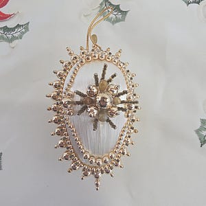May include: An oval-shaped Christmas ornament featuring a white satin-wrapped base. The perimeter is decorated with gold-toned beads, while the center displays a cluster of gold spheres and small brown accents. A gold hanging cord is attached at the top.