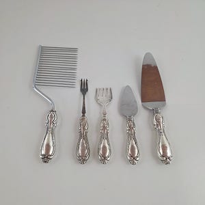 Op de afbeelding: Een set van vijf zilverkleurige serveerbestekken met sierlijke, reliëf handvatten. De collectie bevat een brede snijder met meerdere tanden, een smalle vork met twee tanden, een bredere vork met vier tanden, een klein driehoekig smeermes en een grote driehoekige taartschep.