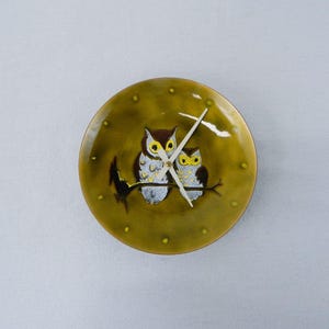 May include: A round wall clock with a painted owl design. The clock face is olive green with two owls perched on a branch. The clock hands are white. The clock is approximately 8 inches in diameter.