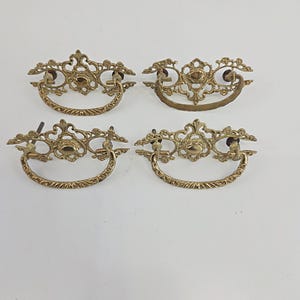 May include: Four antique brass drawer pulls with ornate, filigree designs. Each pull features a decorative center and a curved handle. The brass has a warm, aged patina, suggesting vintage or retro styling. These pulls are suitable for furniture restoration or adding a vintage touch.