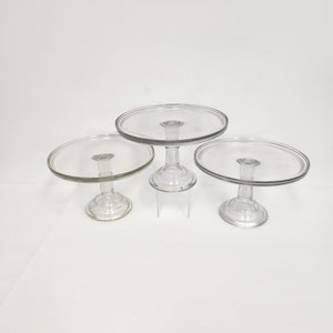 May include: Three clear glass cake stands of varying sizes. The stands have a round base and a round top plate.