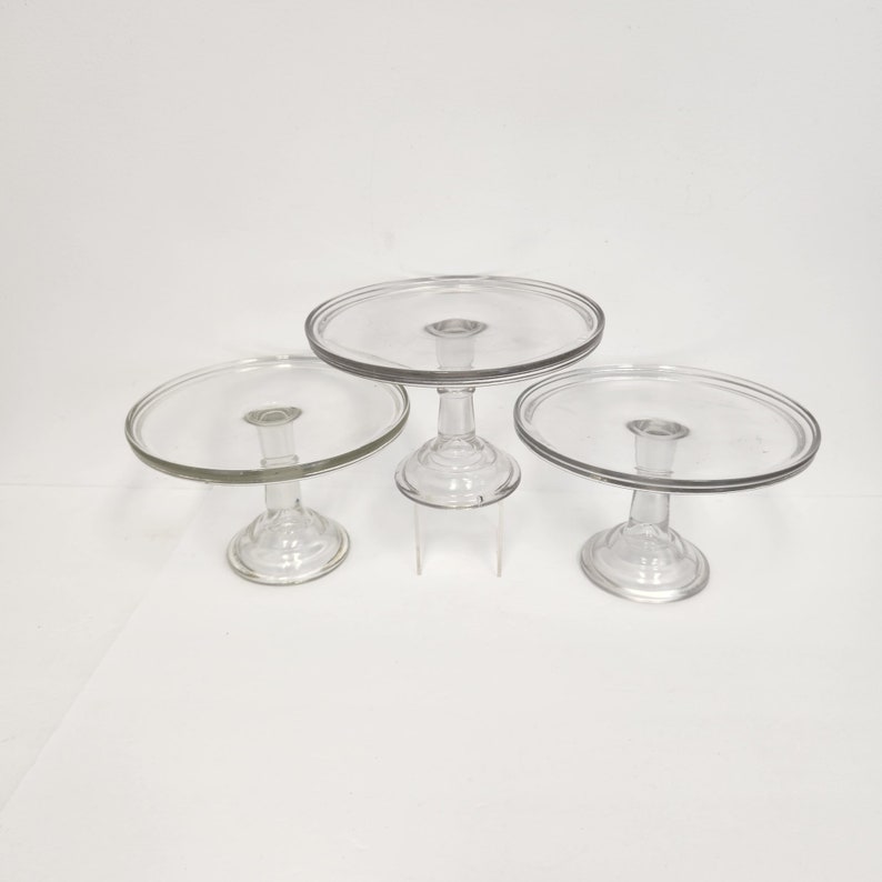 May include: Three clear glass cake stands of varying sizes. The stands have a round base and a round top plate.
