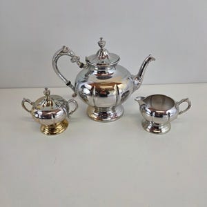 May include: A silver tea set with a teapot, creamer, and sugar bowl. The teapot has a curved handle and a lid with a decorative finial. The sugar bowl has a lid and two small handles. The creamer has a small handle.