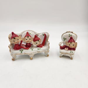 Puede incluir: Tres elfos rojos y blancos sentados en un sofá de porcelana blanca con detalles dorados. Un solo elfo se sienta en una silla de porcelana blanca a juego con detalles dorados.