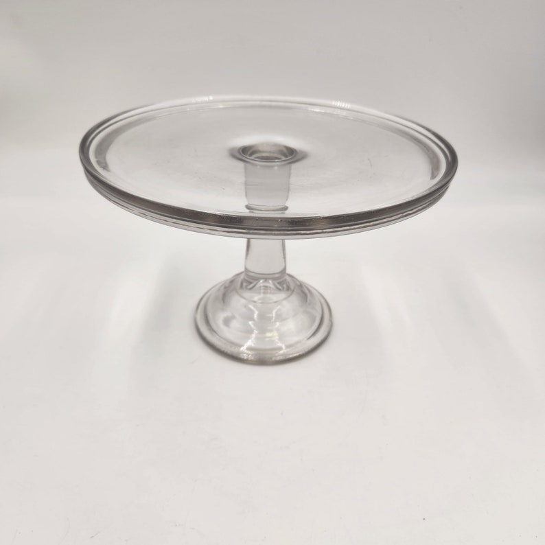 May include: A clear glass cake stand with a round base and a wide, flat top. The stand is perfect for displaying desserts or other treats.
