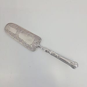 May include: Silver cake server with an ornate design. The server features a rectangular blade with a decorative border and a detailed handle. The handle has a floral design and a curved shape. The server is made of a shiny, reflective metal.