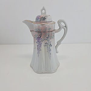 Peut inclure: Une cafetière en porcelaine blanche avec un corps cannelé et un couvercle assorti. La pièce présente un motif peint à la main de fleurs de glycine violettes et de feuillage vert, rehaussé par un liseré doré sur le bord, l'anse et le bec.