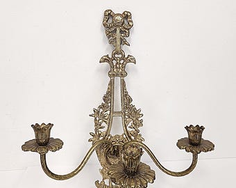 May include: Ornate brass wall sconce with two candle holders. The sconce features a decorative bow, floral accents, and curved arms. The candle holders have a flower-like design. A vintage-style home decor item.