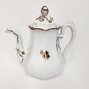 Puede incluir: Tetera de porcelana blanca con ribete dorado y diseño floral. La tetera tiene una tapa con un pomo en la parte superior.