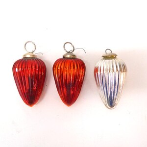 Puede incluir: Tres adornos navideños vintage. Los adornos tienen forma de lágrima con estrías verticales. Dos son de color rojo-naranja y uno plateado con detalles azules. Cada uno tiene una tapa de metal plateado y un lazo de alambre para colgar.