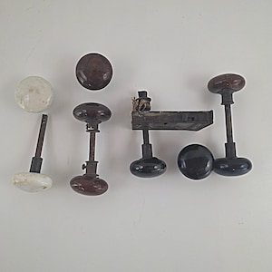 May include: A collection of vintage door hardware including one white ceramic knob, three dark brown mottled knobs, and two black knobs. The set features several metal spindles and a rectangular metal latch mechanism with a small piece of twine attached.