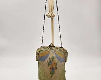 May include: An antique metal mesh handbag with a floral design. The bag features a decorative frame with intricate detailing and a chain handle. The mesh is a light green color, with a colorful floral pattern in the center. The bag is displayed on a decorative stand.
