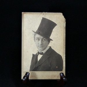 May include: Vintage black and white portrait of a man wearing a top hat and suit. The photograph is mounted on a card with a decorative border. The man is looking directly at the camera.
