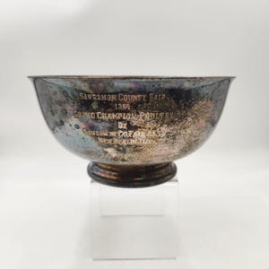 May include: A silver-toned bowl with a dark rim and a pedestal base. The bowl has text that reads "Sangamon County Fair 1955 Grand Champion Poultry". The bowl is displayed on a clear acrylic stand against a white background.